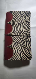 A zip-around journal or notebook cover featuring a black and beige zebra print on the front cover and a deep red leather spine with decorative rivets, held together by a black elastic band.