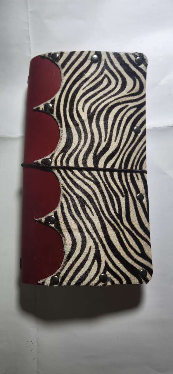 A zip-around journal or notebook cover featuring a black and beige zebra print on the front cover and a deep red leather spine with decorative rivets, held together by a black elastic band.