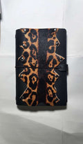 A black A5 leather journal with a leopard print pattern on the front cover, secured with a black elastic band and strap closure, resting on a white sheet of paper, desgined by Mdame Kim in South Korea