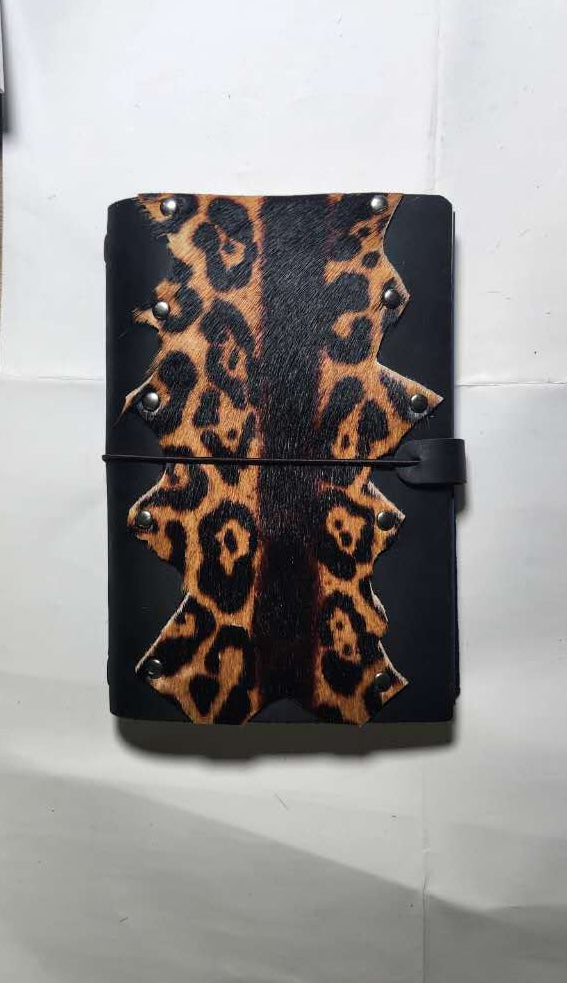 A black A5 leather journal with a leopard print pattern on the front cover, secured with a black elastic band and strap closure, resting on a white sheet of paper, desgined by Mdame Kim in South Korea