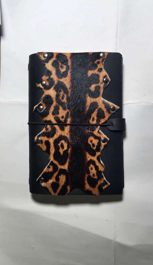A black A5 leather journal with a leopard print pattern on the front cover, secured with a black elastic band and strap closure, resting on a white sheet of paper, desgined by Mdame Kim in South Korea